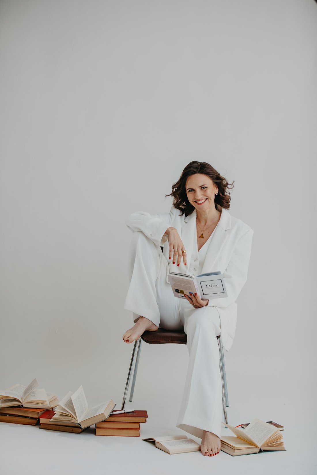 Katharina Pohl im weißen Anzug auf einem Stuhl mit Buch, lächelnd
